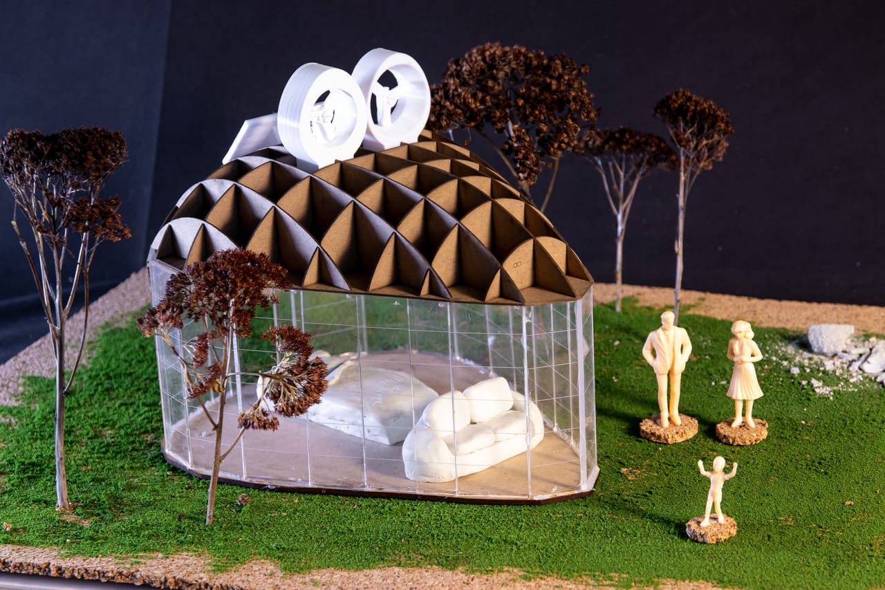 Physical model of a small glass pavilion with a ribbed brown dome roof and two white fan units on top, a sofa inside, and small trees and figures on green grass