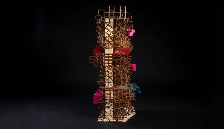 Photograph of a tall model tower made of a dense lattice of thin timber-coloured members on a pale base, with pink, red and blue volumes hanging off its sides, against a black background