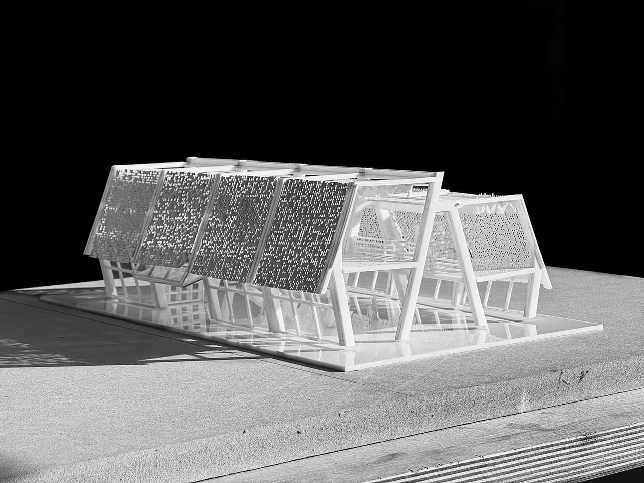 Black-and-white photograph of a white physical model of a long building made of repeated A-shaped frames, with perforated panels on the sloping roof faces, on a pale base against a black background