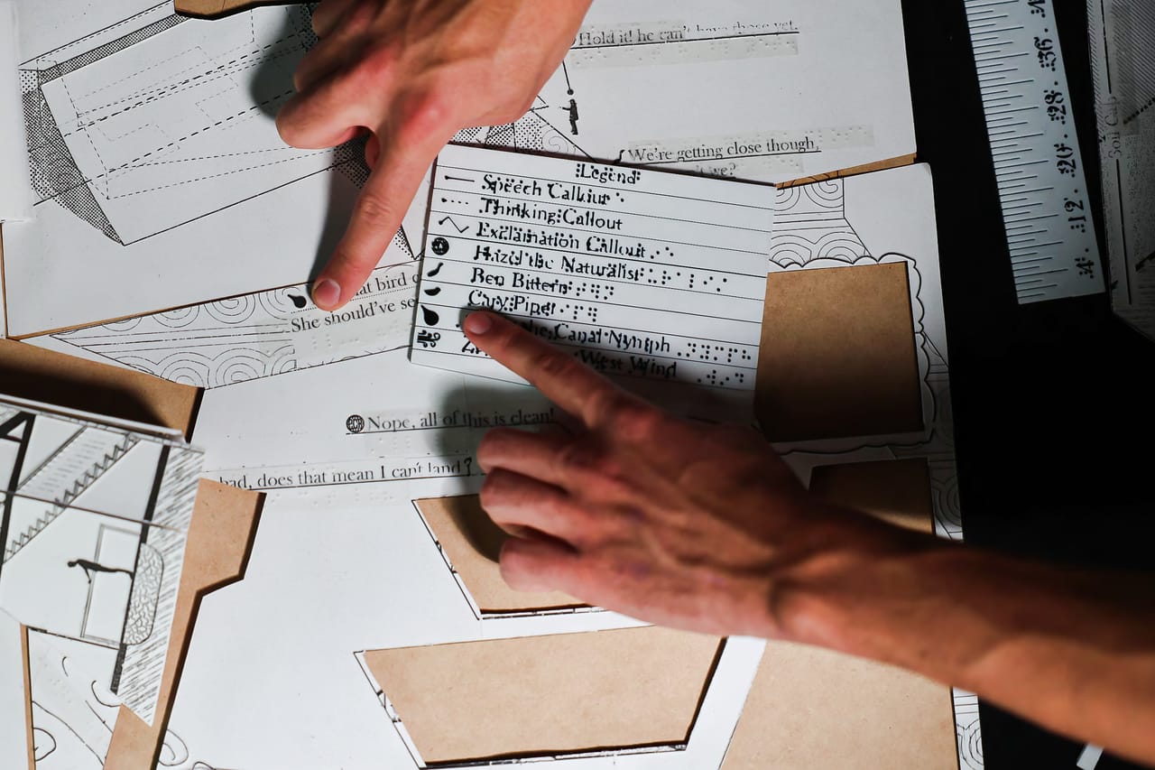 Seen from above, one hand points at a card of lettering lying on a white paper-and-card model while another hand rests on a sheet of drawings. A ruler stands at the right edge.