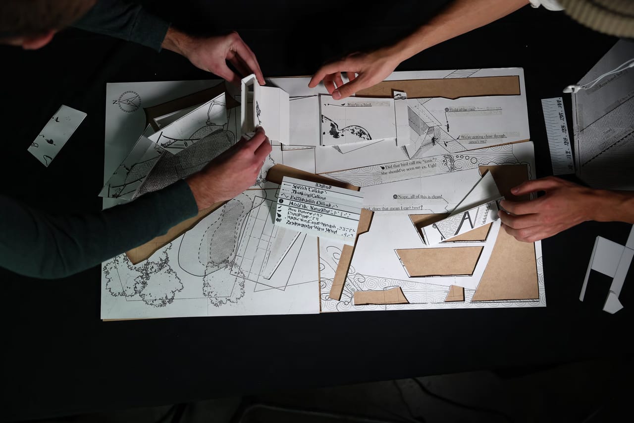 Seen from above, several hands work over a white paper-and-card model on a black table.