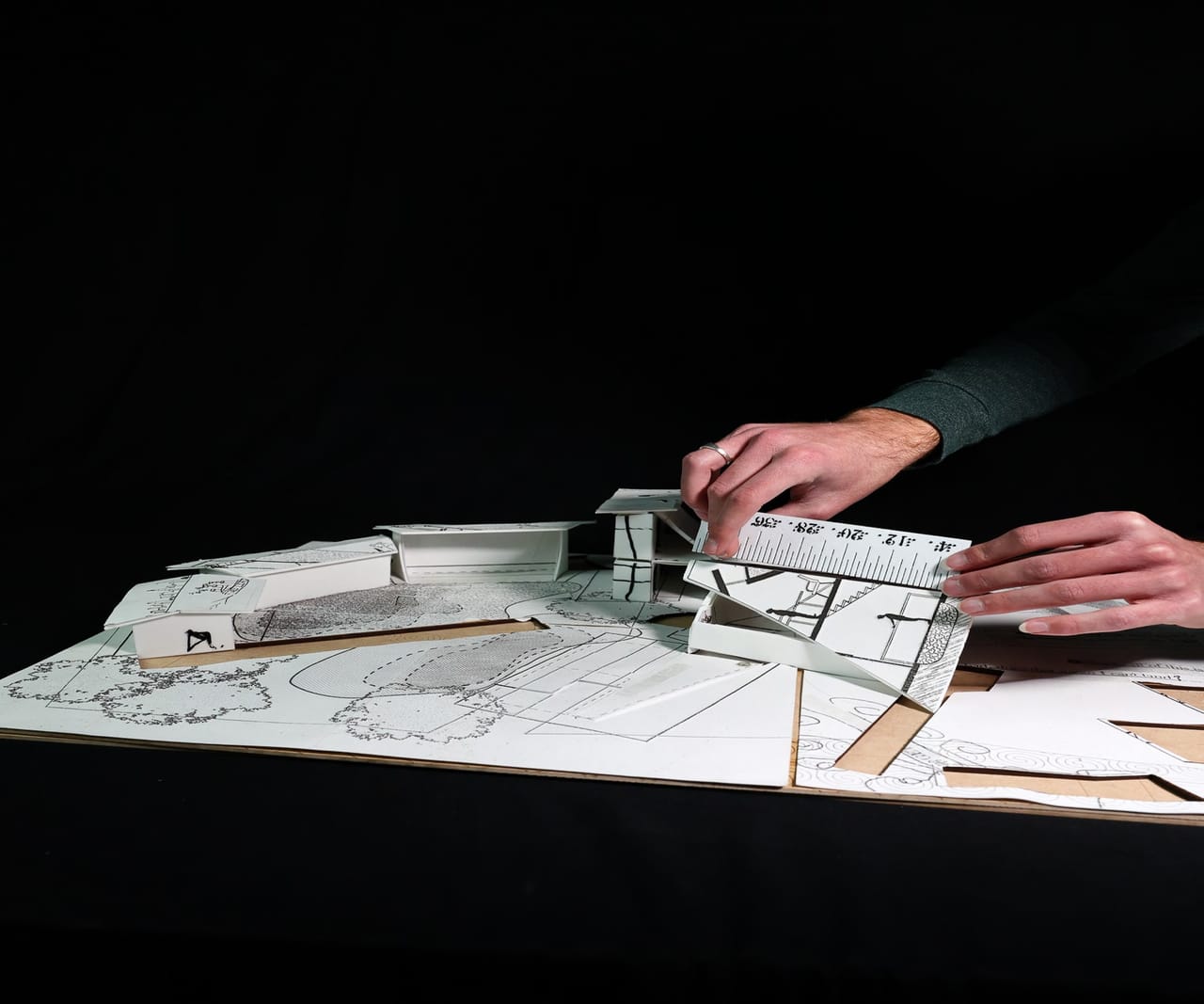 A white paper-and-card model with raised pieces on a black table. A hand rests at the right edge.